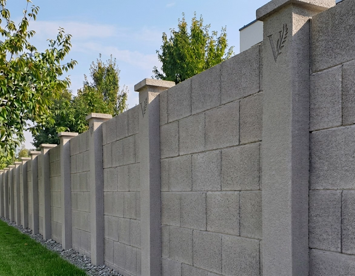 Precast concrete block perimeter wall running along a green grass lawn under a clear blue sky. The wall features a clean, minimalist "V" logo with a stylized leaf branch neatly etched directly into the concrete surface.