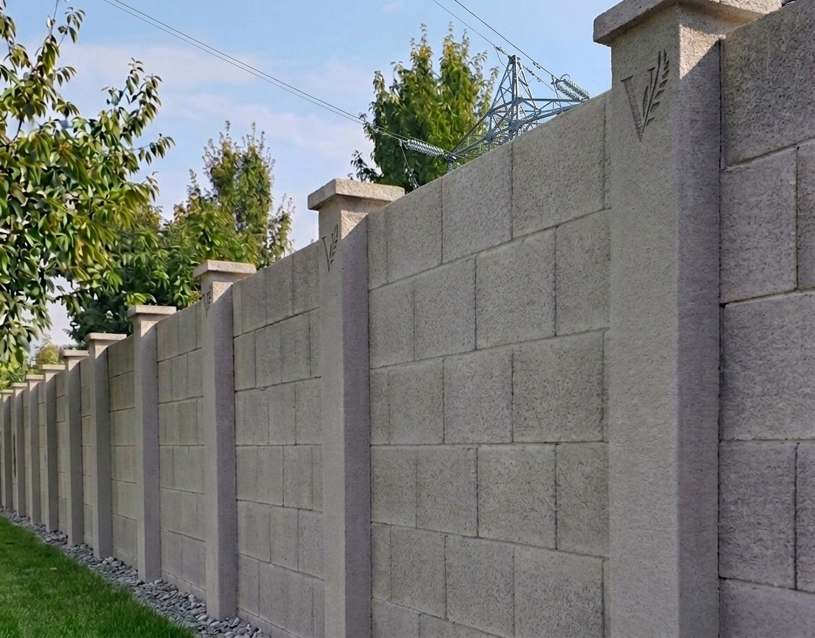 Precast concrete fence wall with column system and textured panels running along a landscaped lawn, with utility transmission tower and power lines visible in the background behind residential trees.