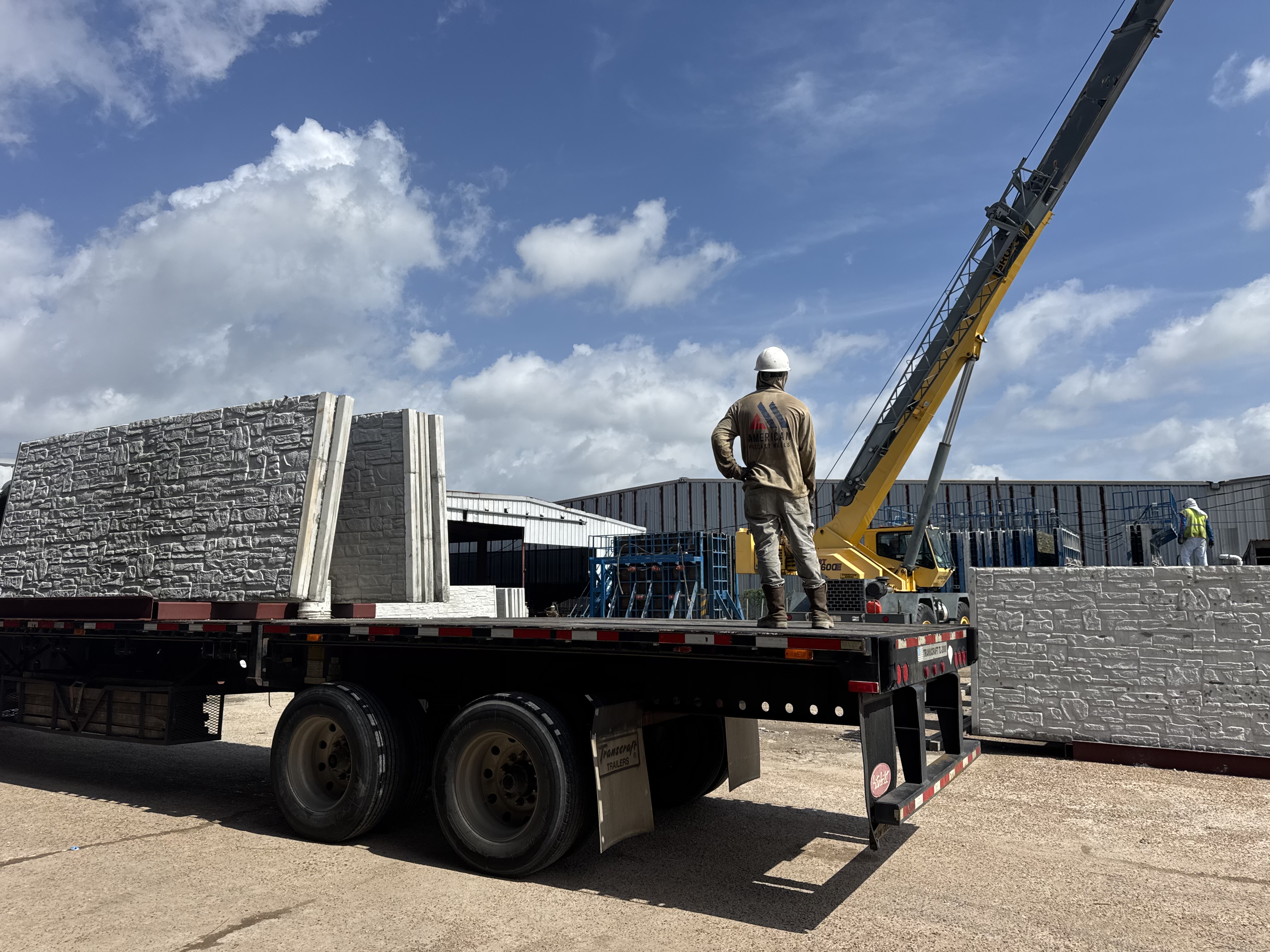 A construction worker in a hard hat and American Precast Walls shirt stands on a flatbed trailer, overseeing the loading of large, textured stone-look concrete wall panels by a mobile crane on a sunny day.