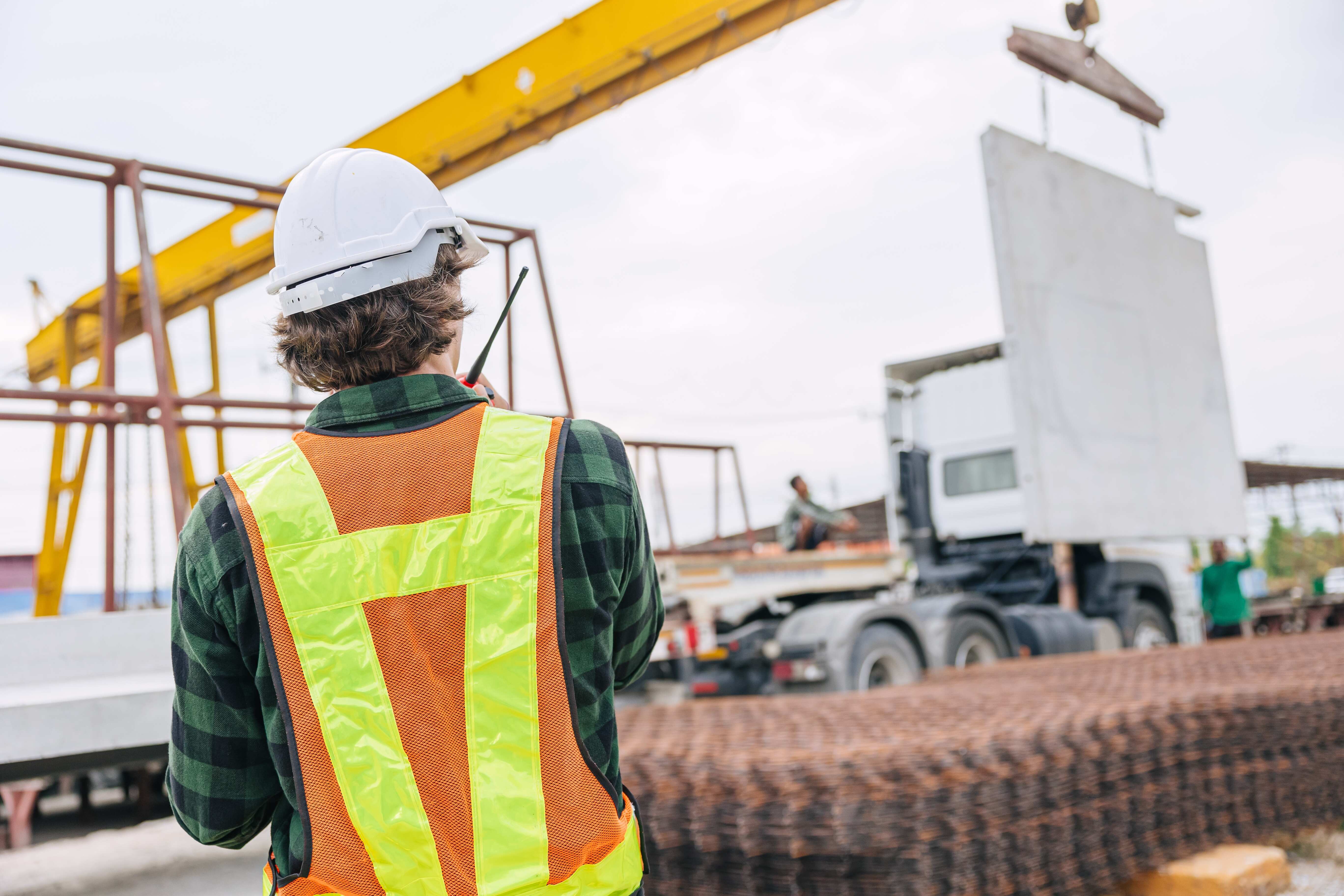 Construction site supervisor in high-visibility vest and hard hat using a walkie-talkie to coordinate the crane lifting of a precast concrete wall panel