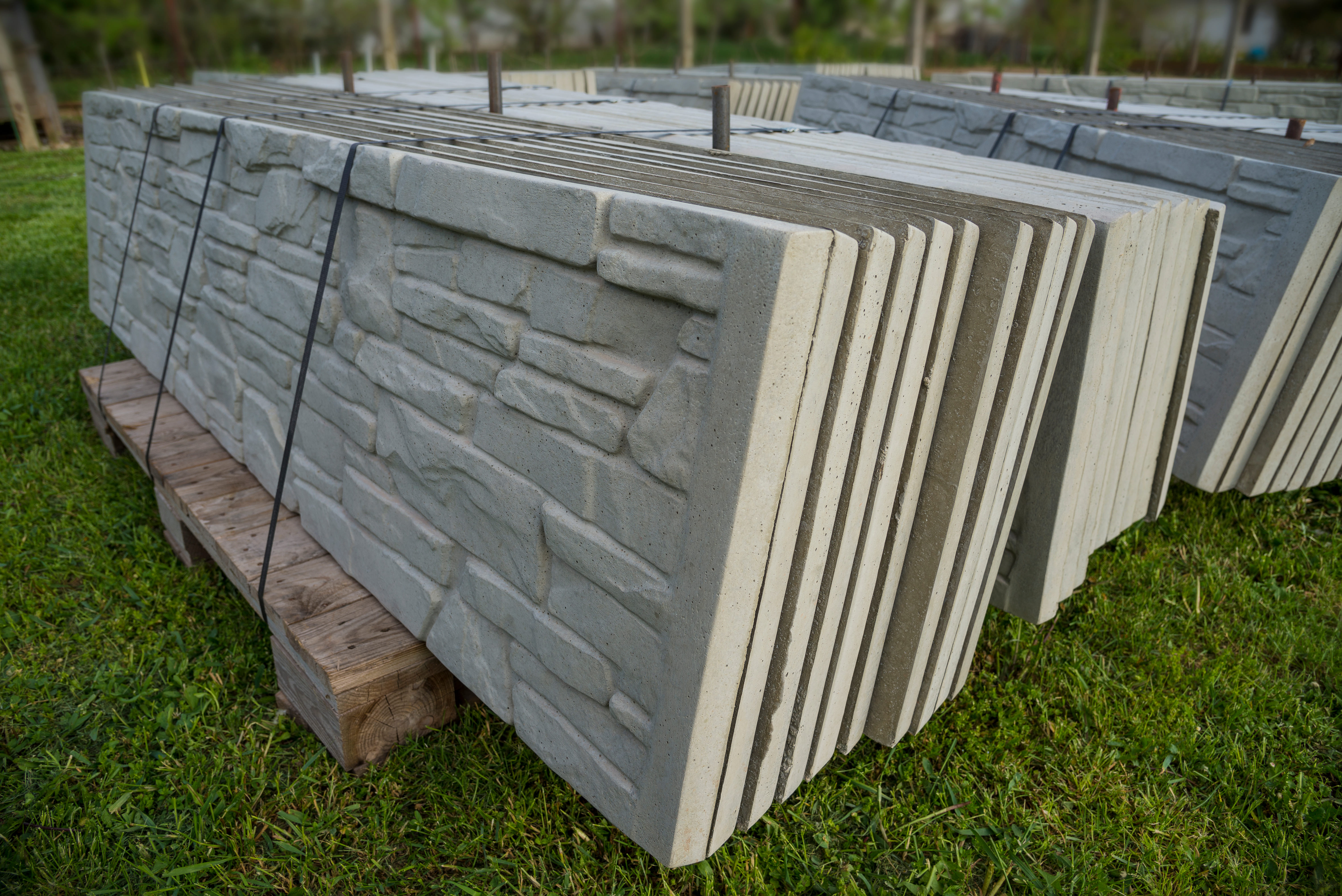 A stack of precast concrete fence panels with a textured stone-look finish resting on a wooden pallet in a grassy outdoor area