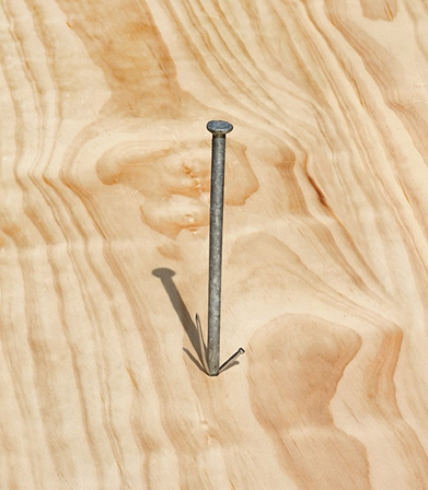 Close-up of a long metal nail driven upright into a light wooden surface casting a shadow.