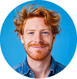 Smiling red-haired man with curly hair wearing a denim jacket against a blue background.