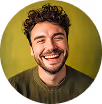 Smiling man with curly hair and beard wearing a dark green shirt against a yellow background.