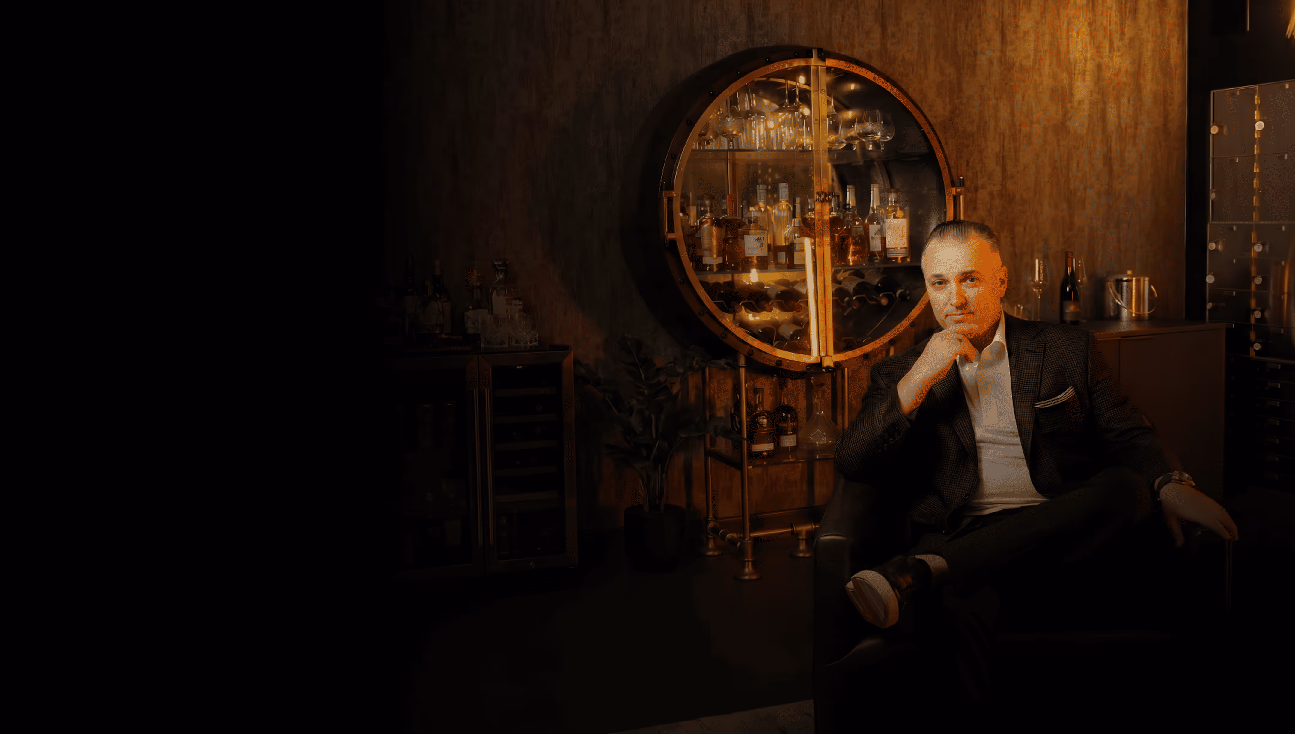 Man in a blazer sitting thoughtfully in a dimly lit room with a circular cabinet of liquor bottles behind him.
