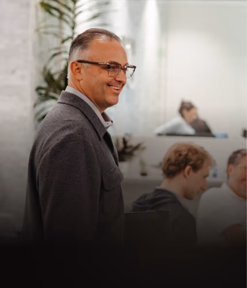 Smiling middle-aged man wearing glasses and a gray jacket standing in an office with blurred colleagues in the background.