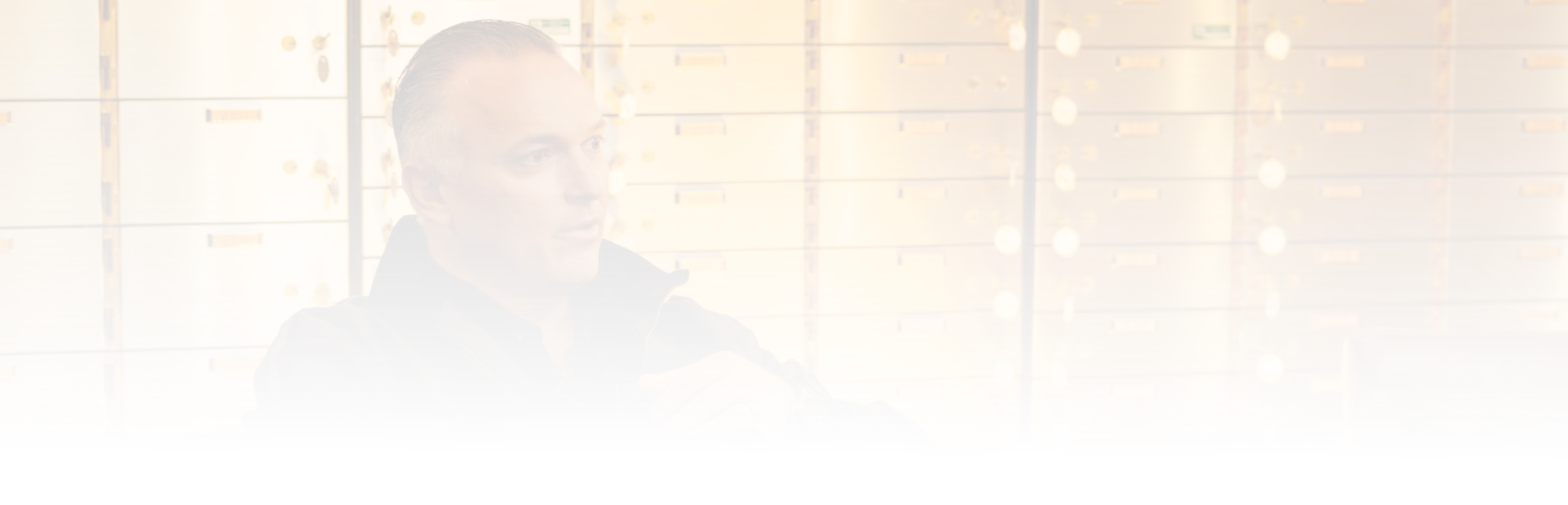 Man with slicked-back hair wearing a dark jacket, sitting and gesturing with his hands in front of gold-colored safety deposit boxes.