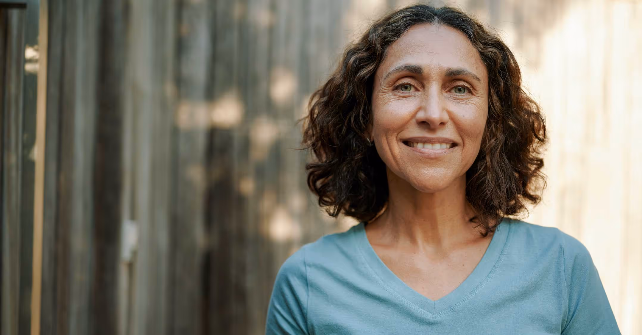 Smiling middle-aged woman with curly hair wearing a light blue V-neck shirt standing outdoors with a wooden background.
