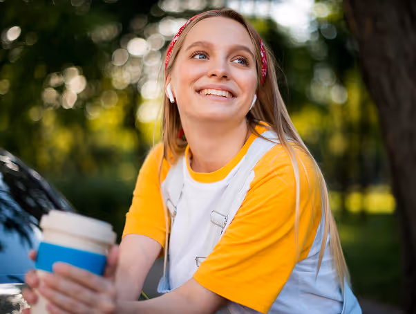 Smiling young woman wearing a yellow and white shirt with a red headband and wireless earbuds, holding a disposable coffee cup outdoors.