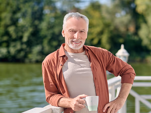 Smiling elderly man with gray hair and beard holding a white mug while leaning on a white railing by a lake surrounded by trees.