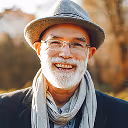 Smiling elderly man with white beard wearing a gray hat, glasses, and a scarf outdoors.