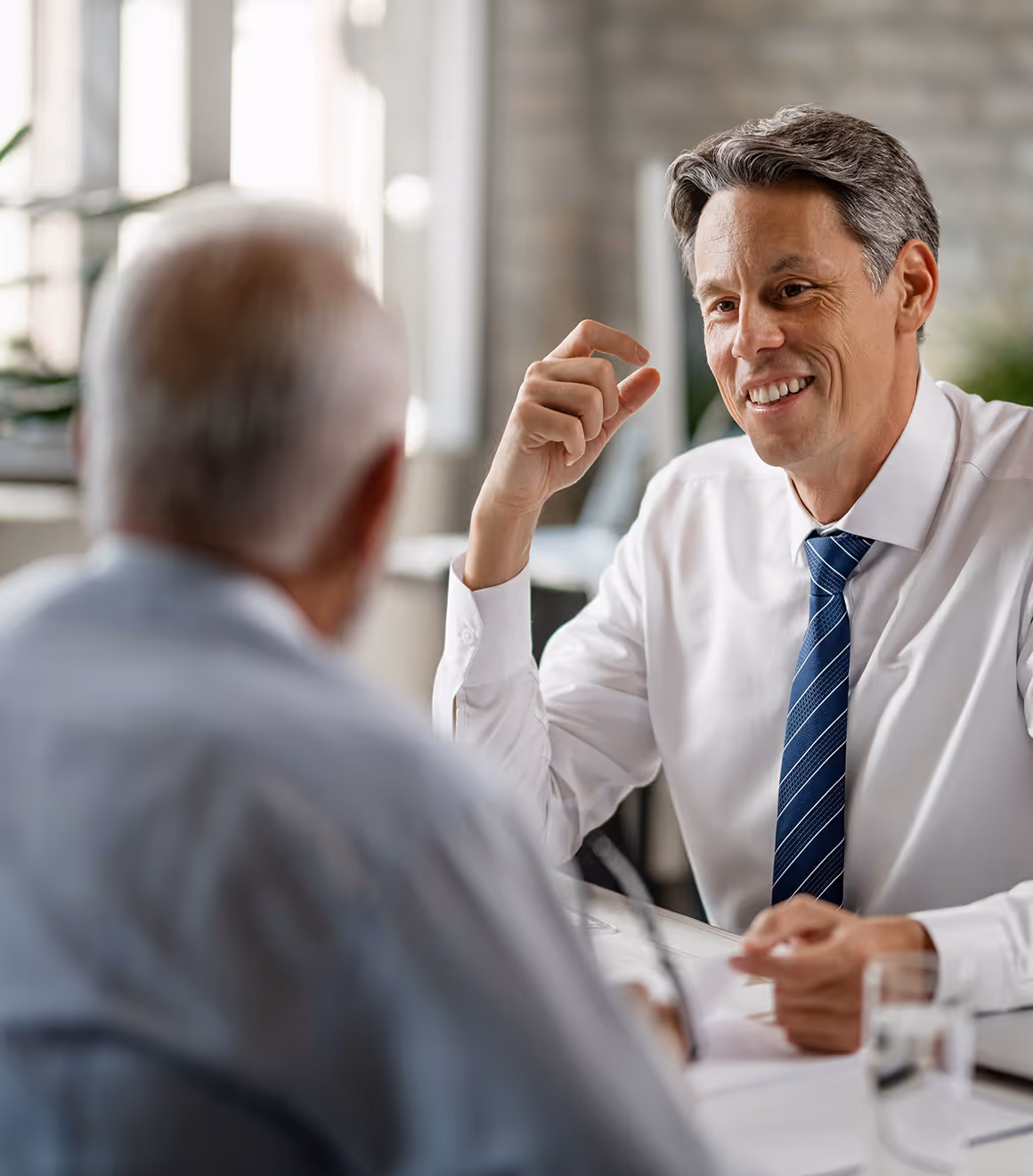 Man in a white shirt and blue striped tie smiling and talking to an older man at a table.