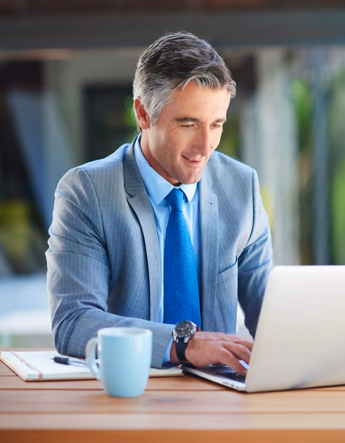 Middle-aged man in a gray suit and blue tie working on a laptop at a wooden table with a blue mug and notebook.