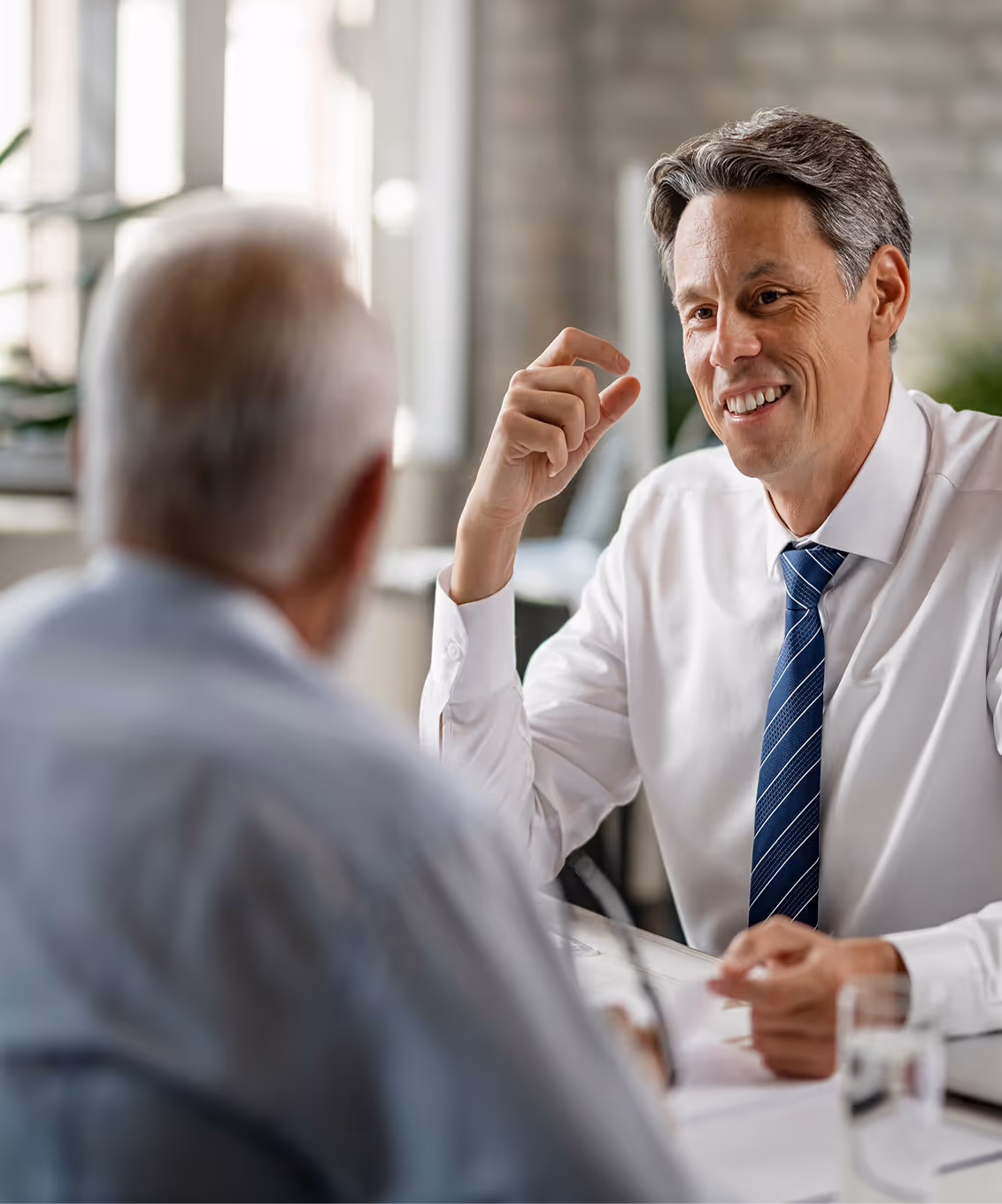 Smiling businessman in white shirt and blue striped tie talking with an older man in a professional office setting.