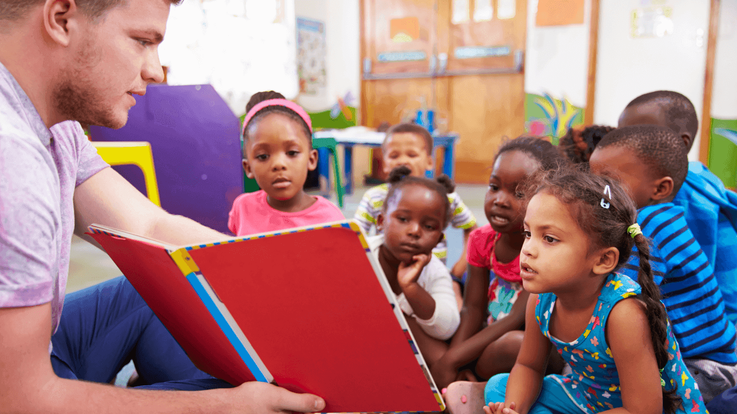 Man reading a large book to a diverse group of young children sitting and listening attentively.