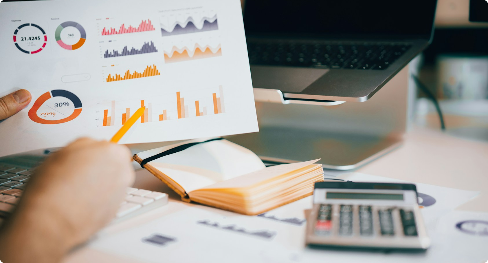 Person pointing at printed charts and graphs with a pencil next to calculator and laptop on desk.
