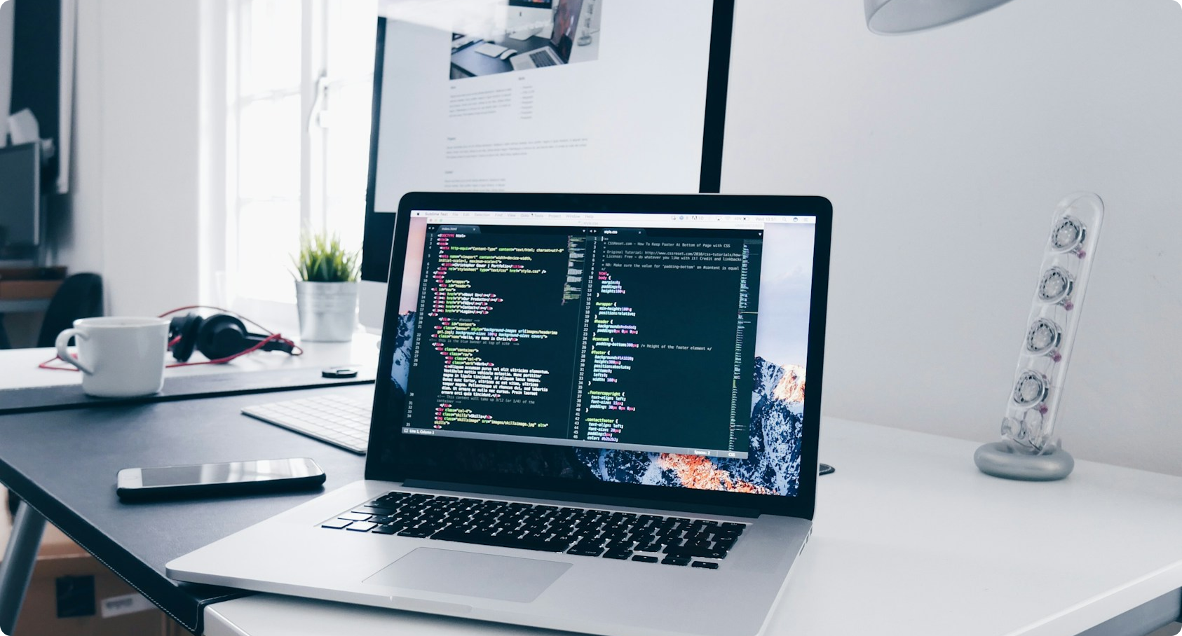 Laptop displaying code editor with HTML and CSS code on a desk with a phone, mug, headphones, and monitor.