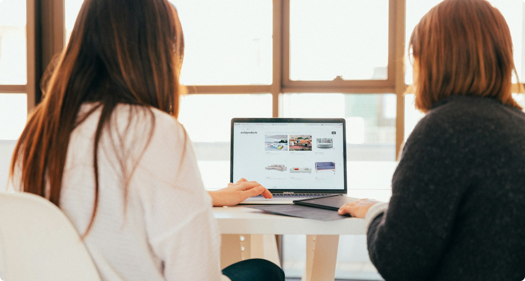 Two women sitting at a table looking at furniture options on a laptop screen.