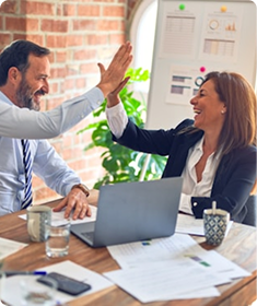 Man and woman in office exchanging a high five over a table with laptop and documents.