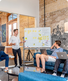 Man presents ideas on whiteboard with sticky notes while two seated men listen and use laptops.