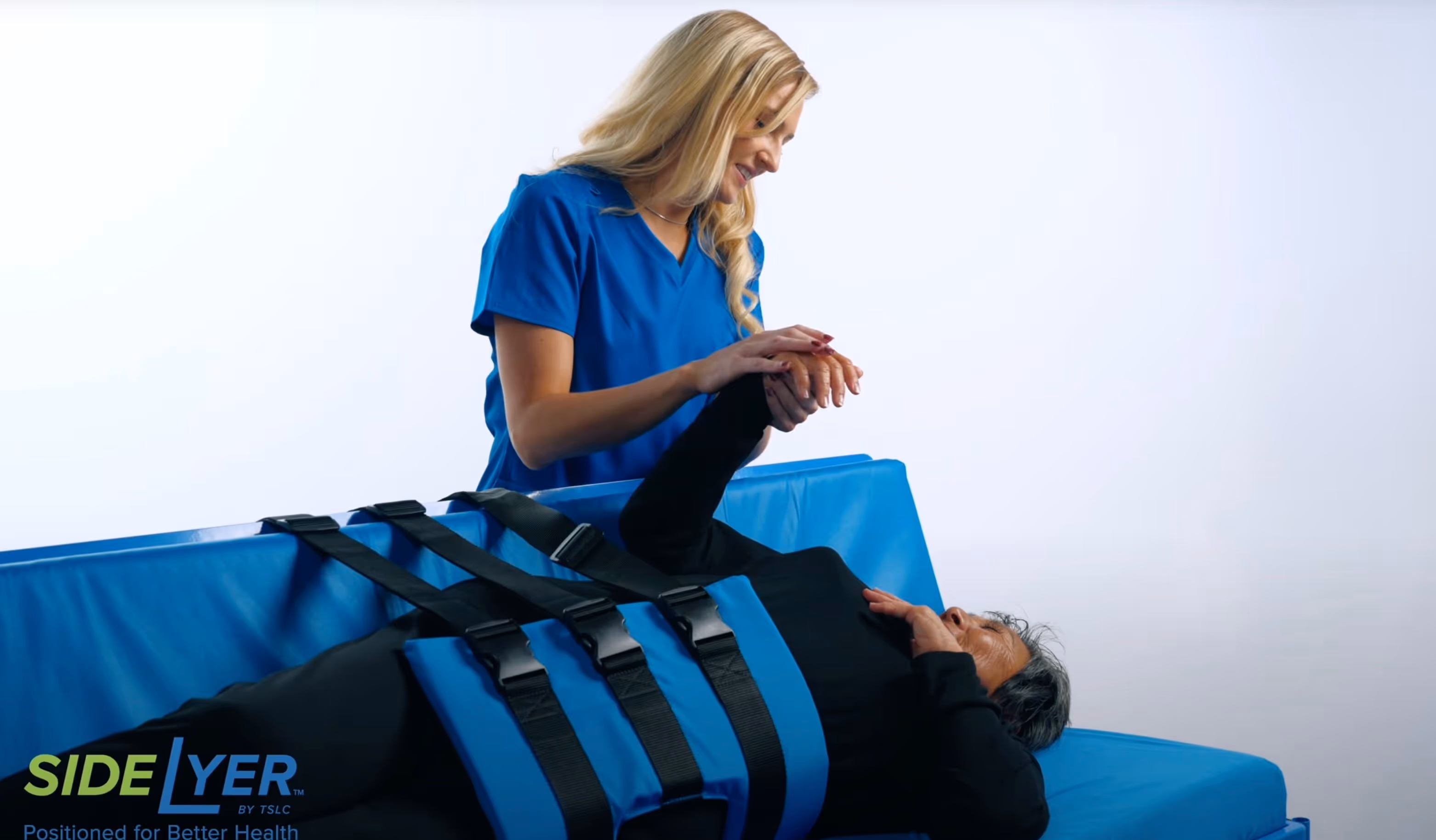 A healthcare professional in blue scrubs assisting an elderly person lying on a blue medical positioning device with black straps.