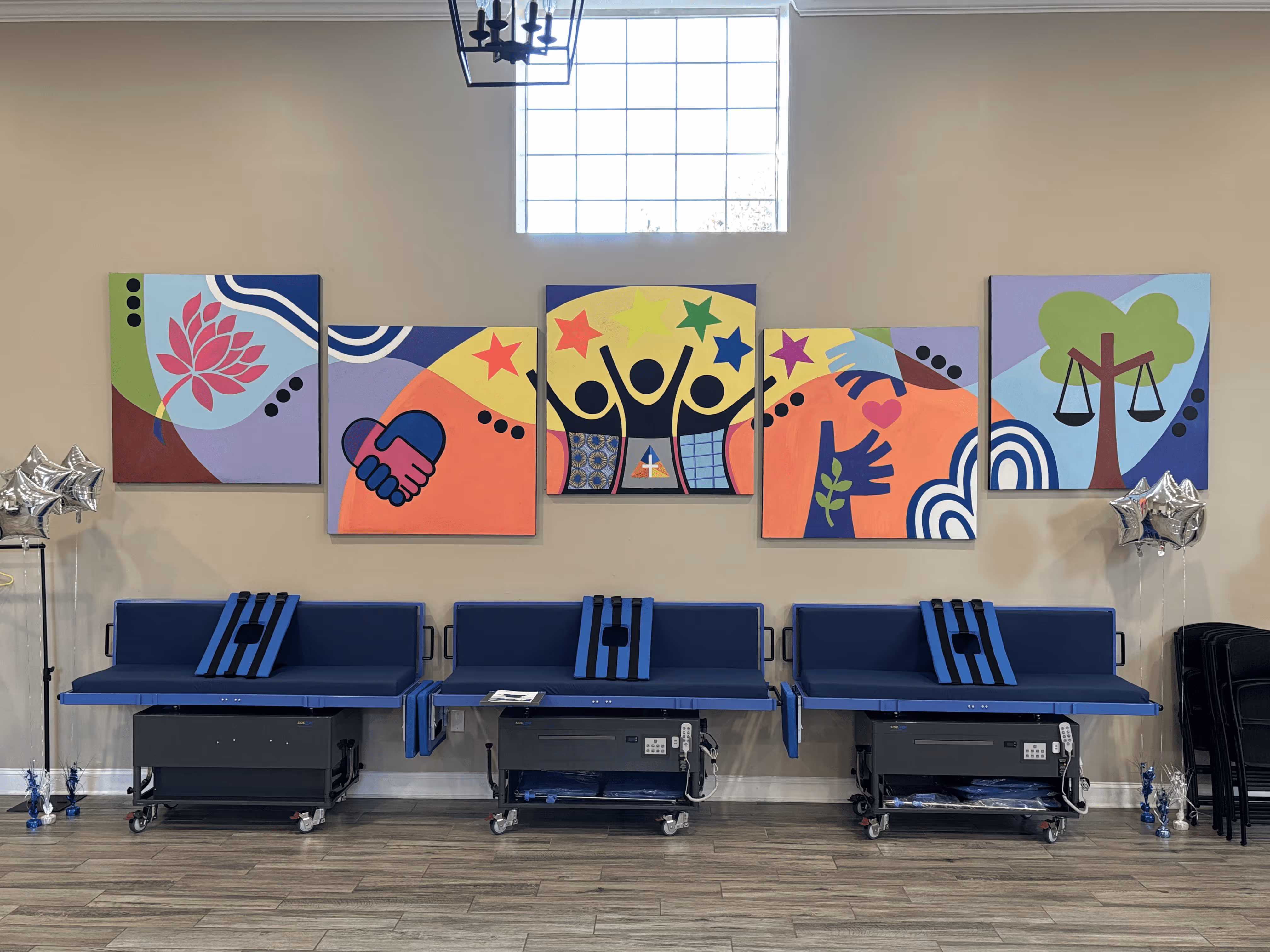 Room with three blue stretchers against a beige wall, colorful abstract paintings above depicting themes of community and justice, and silver star-shaped balloons on either side.