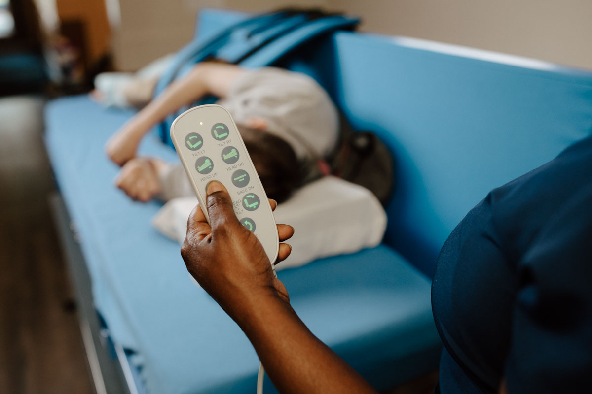 Person holding a remote control for adjusting a hospital bed with a patient lying on the bed in the background.