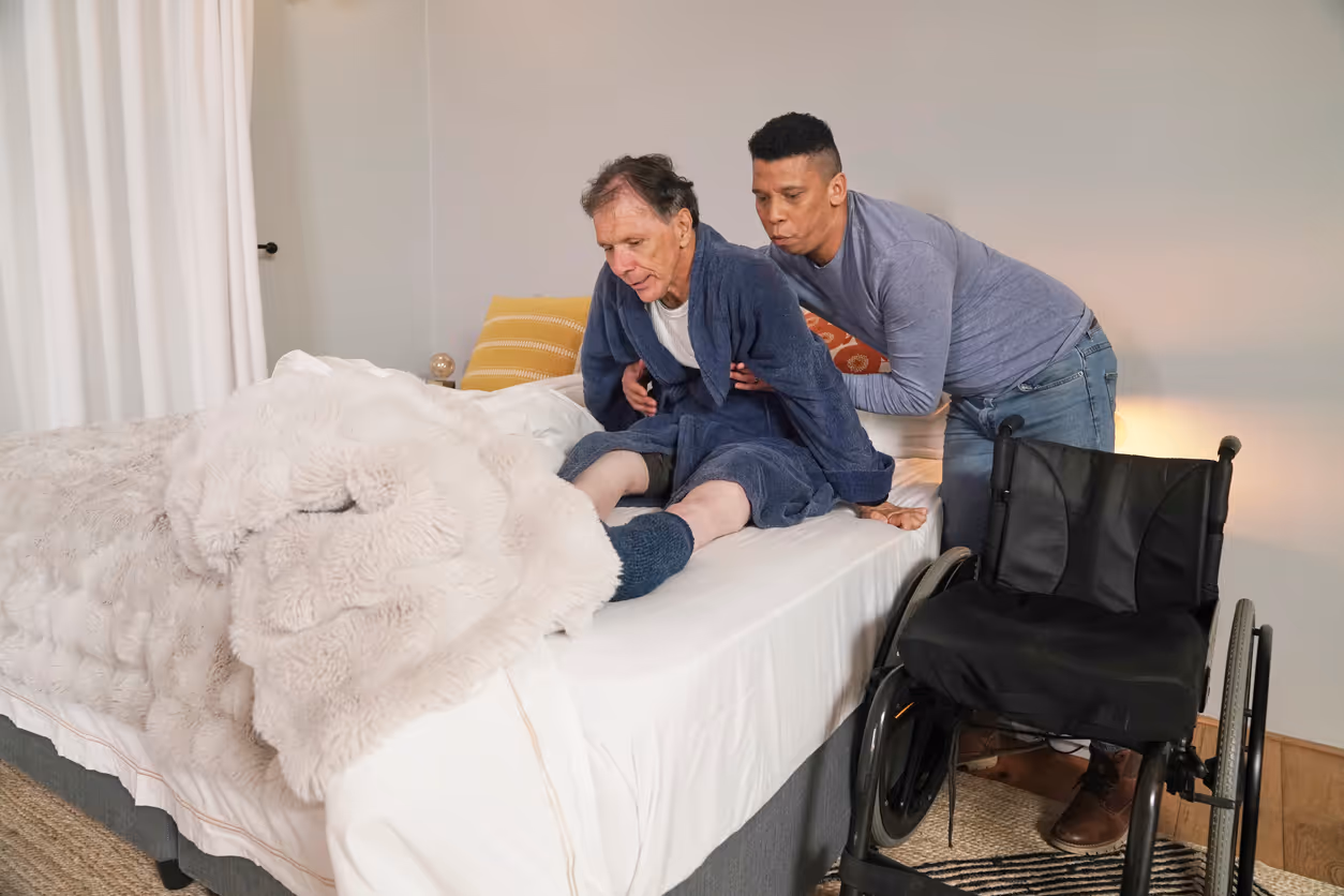 Caregiver assisting an elderly man sitting on a bed next to a wheelchair in a bedroom.