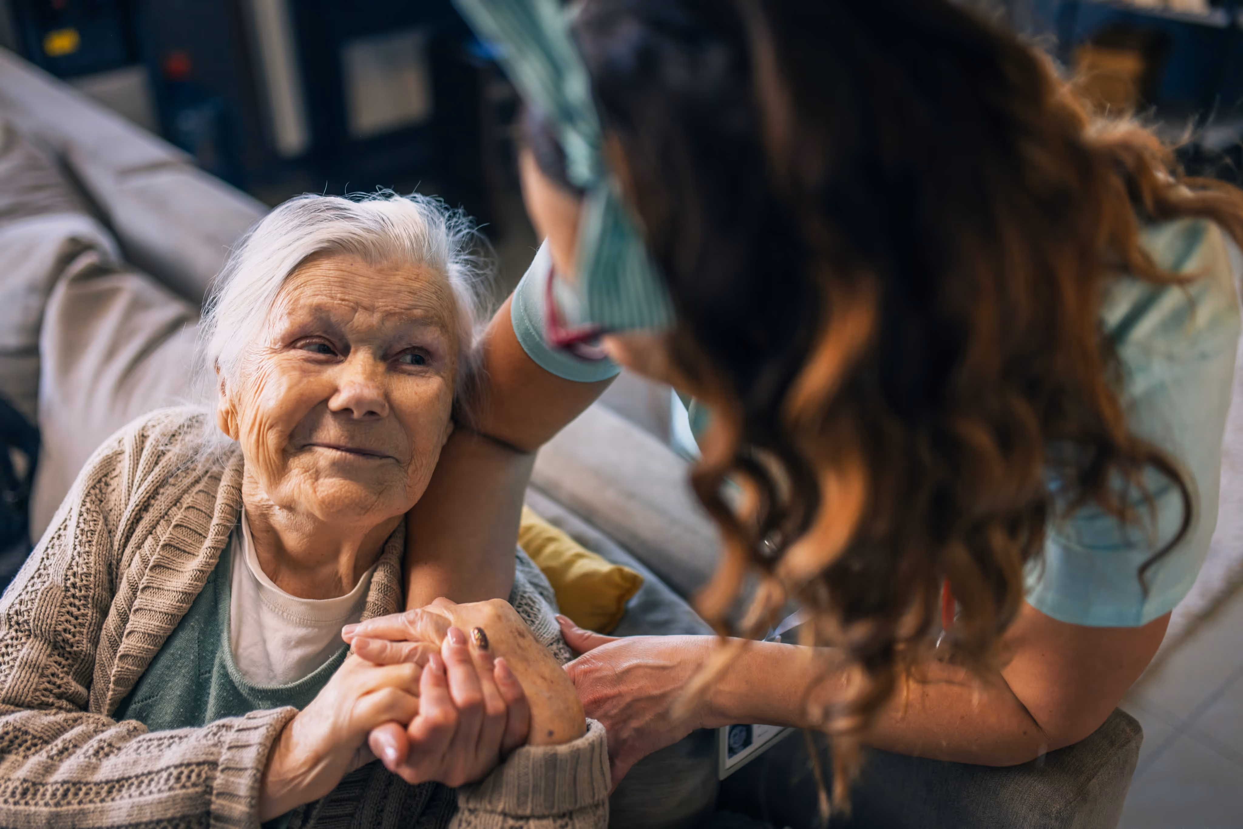 Elderly woman sitting on a couch holding hands with a caregiver who leans toward her.