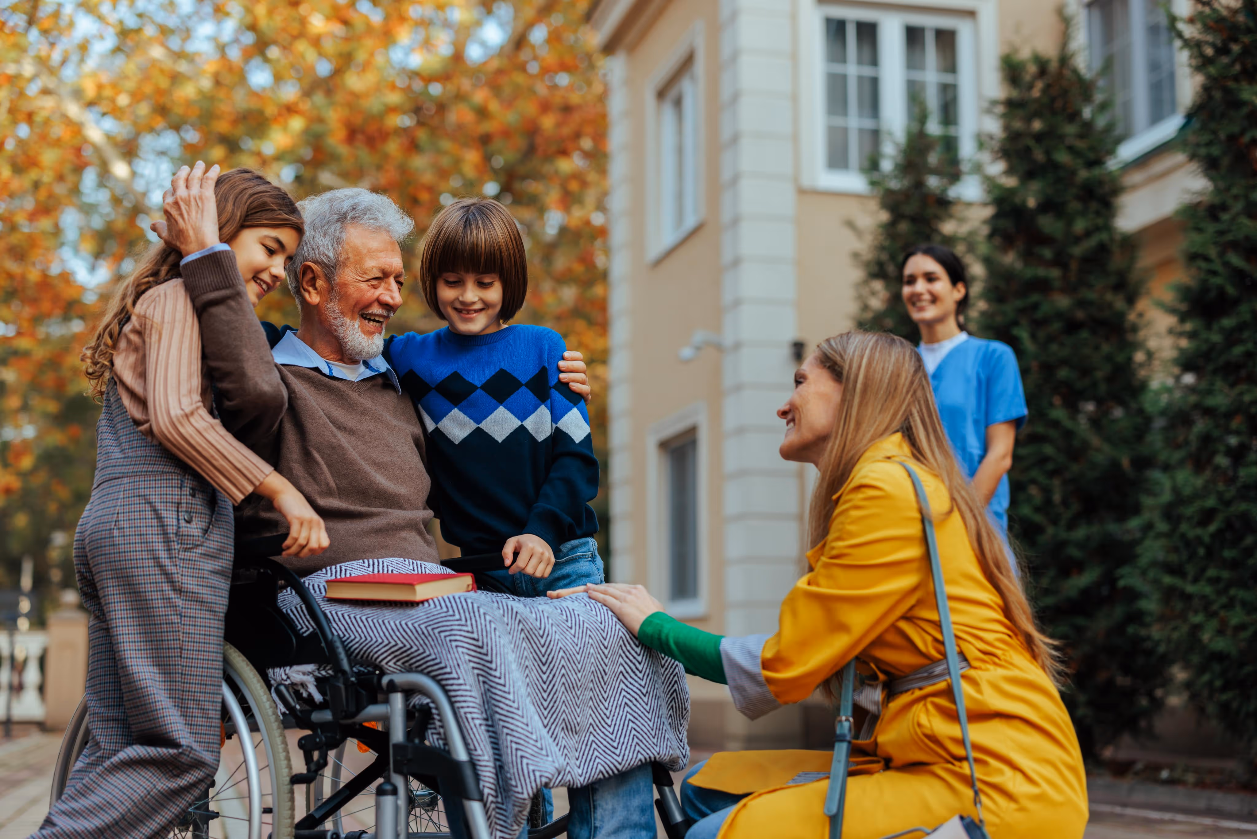 Smiling elderly man in a wheelchair with two children hugging him and a woman in a yellow coat kneeling beside them outdoors.