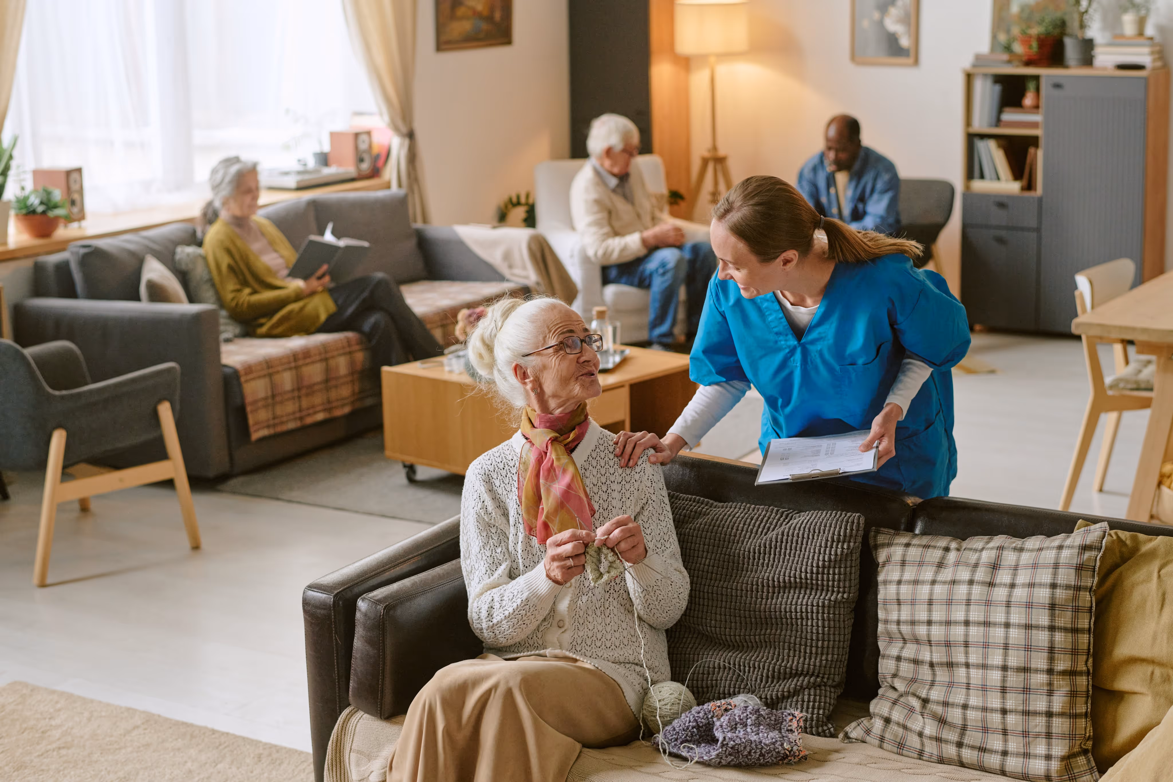 Caregiver in blue scrubs talking to an elderly woman knitting on a couch in a cozy living room with other seniors relaxing in the background.