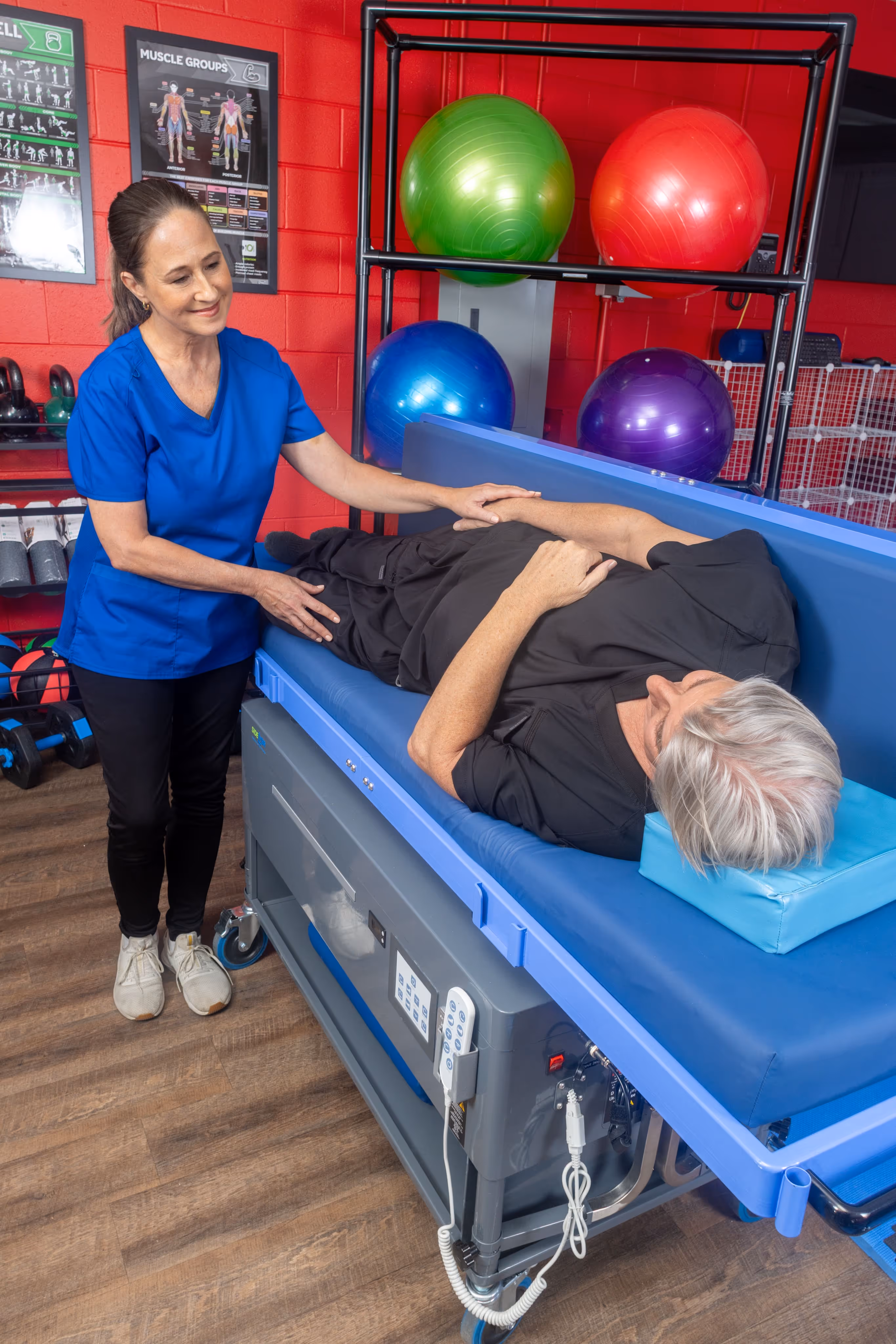 Physical therapist assisting an elderly woman lying on her side on a treatment table in a gym with exercise balls and posters on the wall.