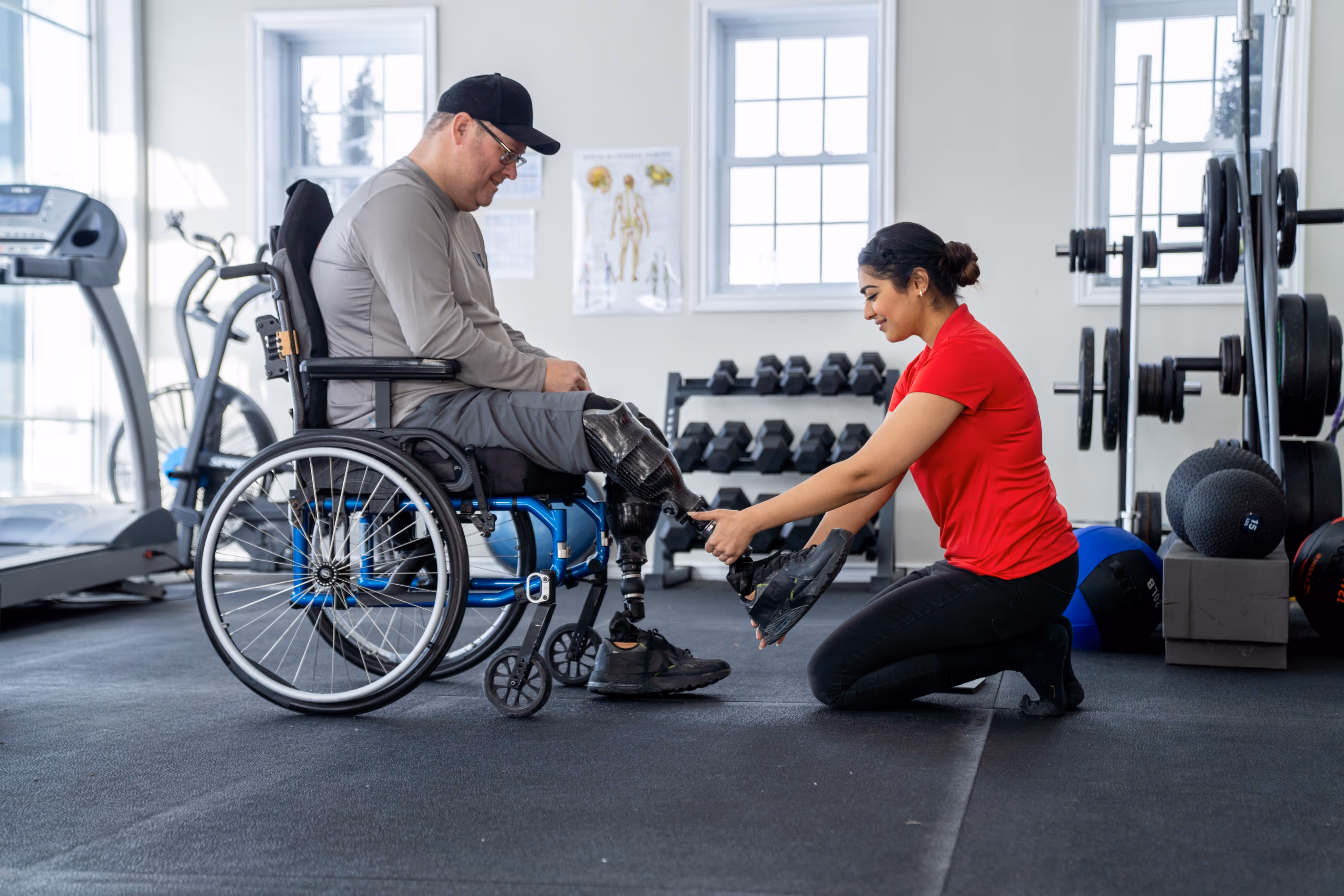 A woman helping a man in a wheelchair by putting on his prosthetic leg shoe in a gym setting.