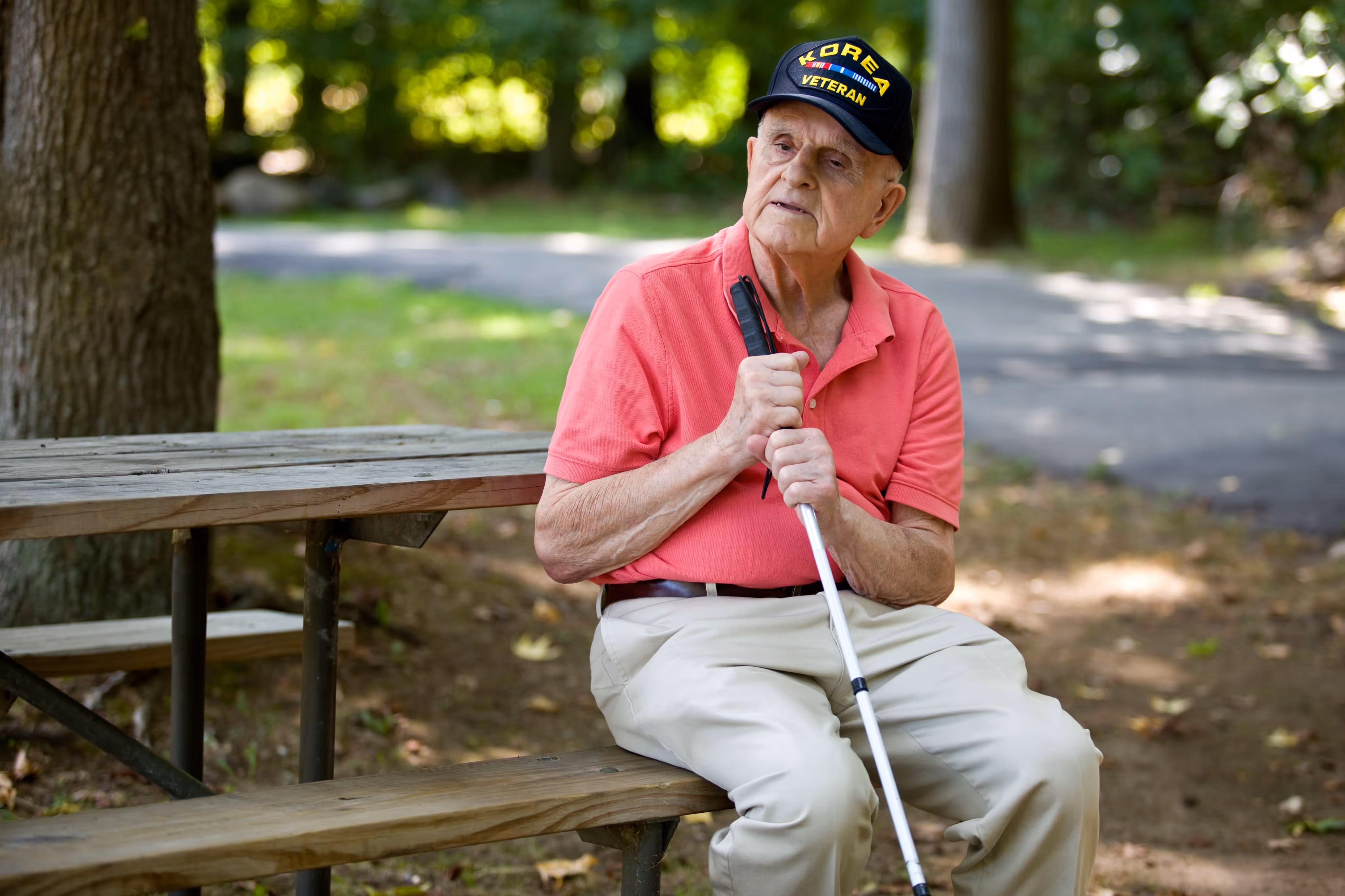 Elderly man wearing a Korea Veteran cap sitting on a wooden bench outdoors holding a white cane.