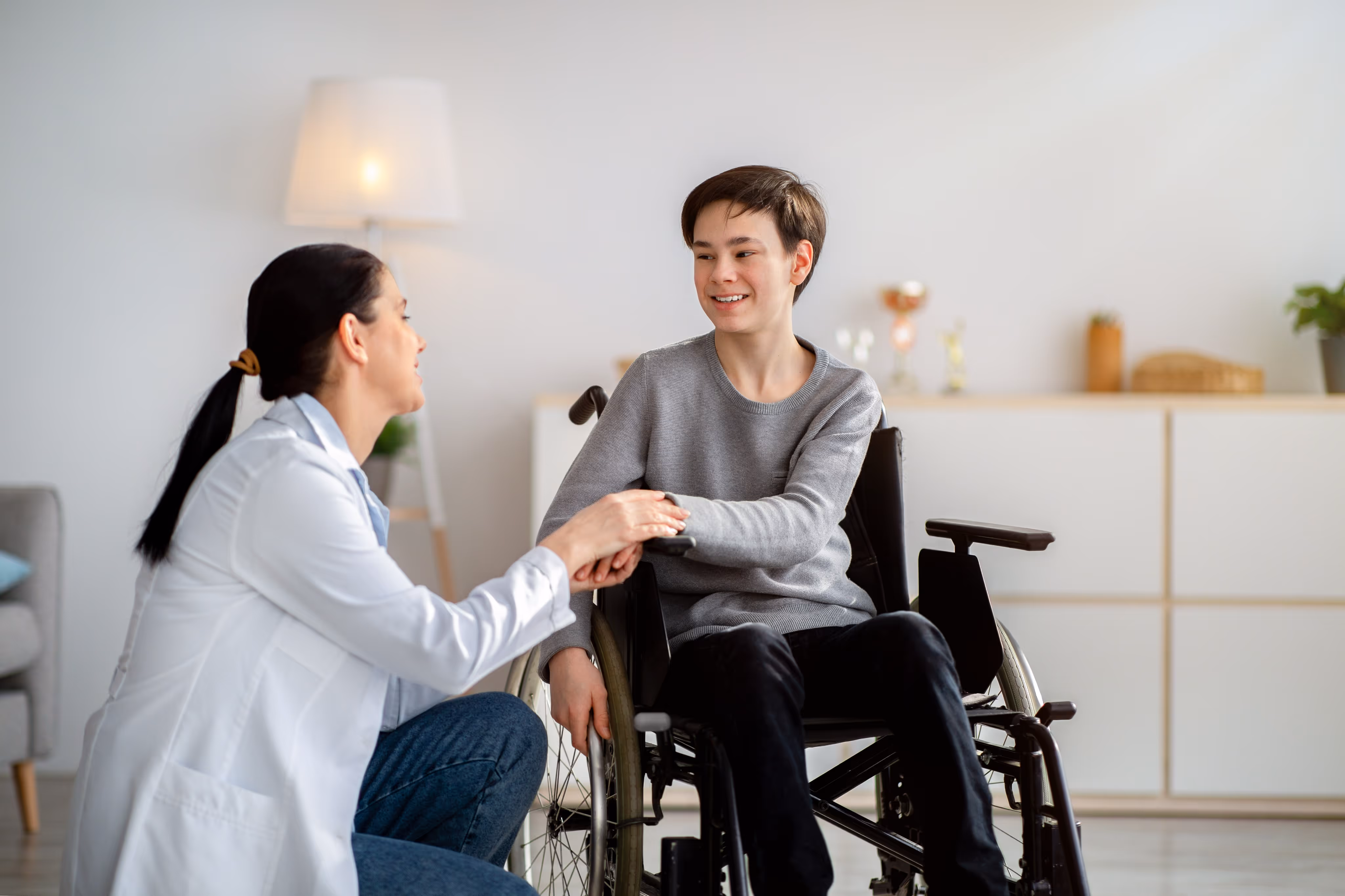 Smiling young person in a wheelchair holding hands with a female doctor in a white coat kneeling beside them.