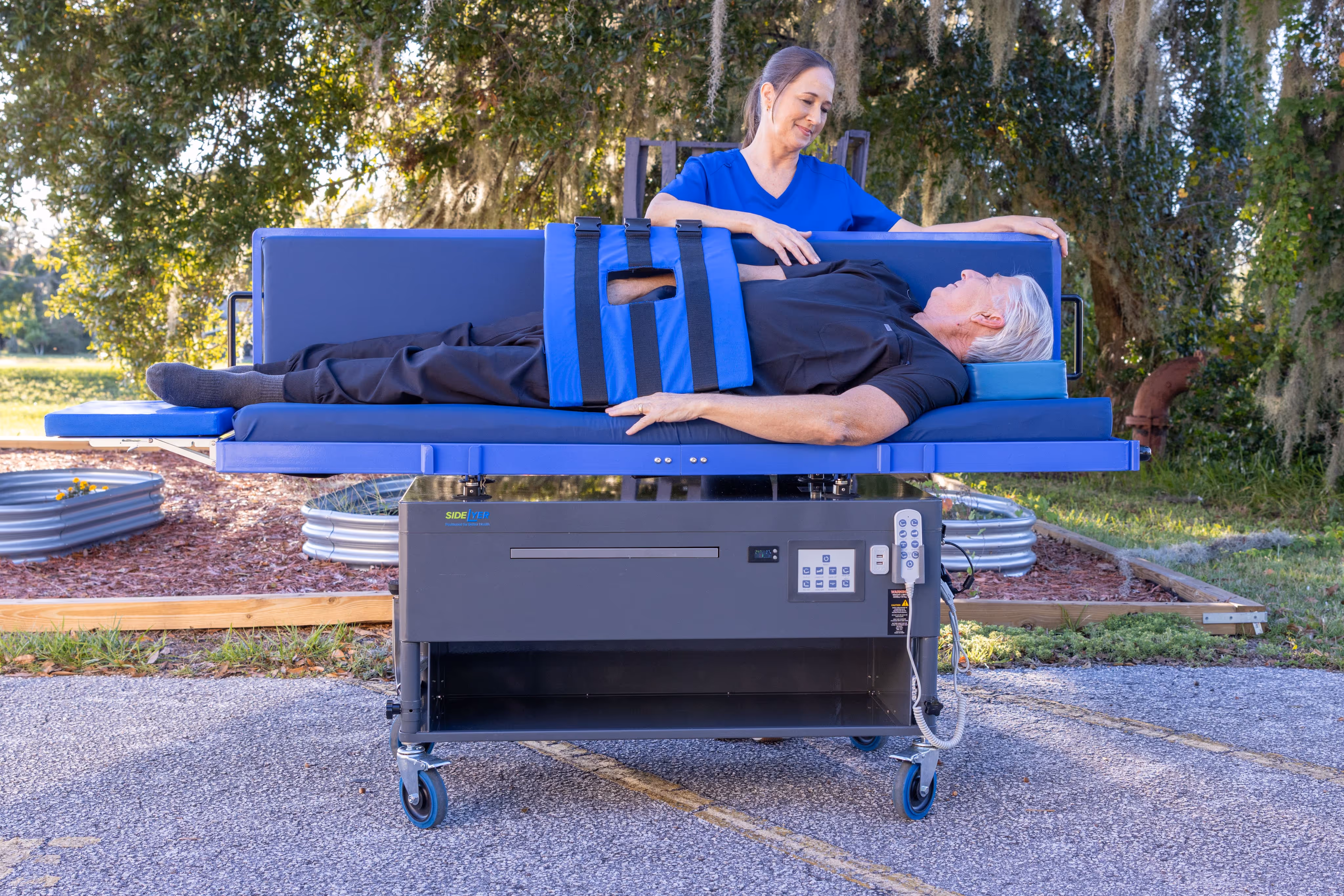 Elderly man lying secured on a blue therapy table outdoors while a female caregiver in blue scrubs stands beside him.