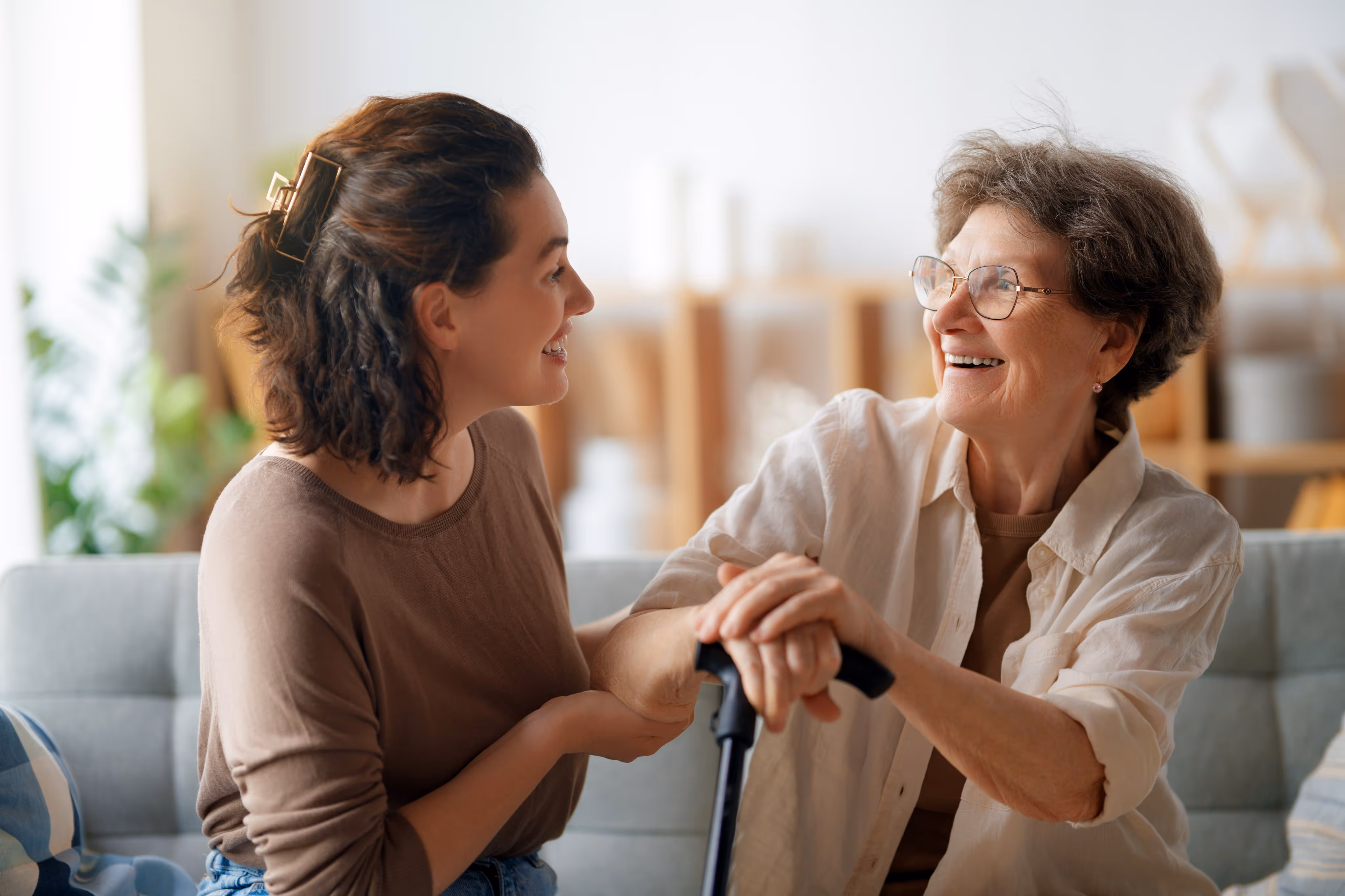 Younger woman smiling and holding the arm of an elderly woman sitting with a cane, sharing a happy moment indoors.