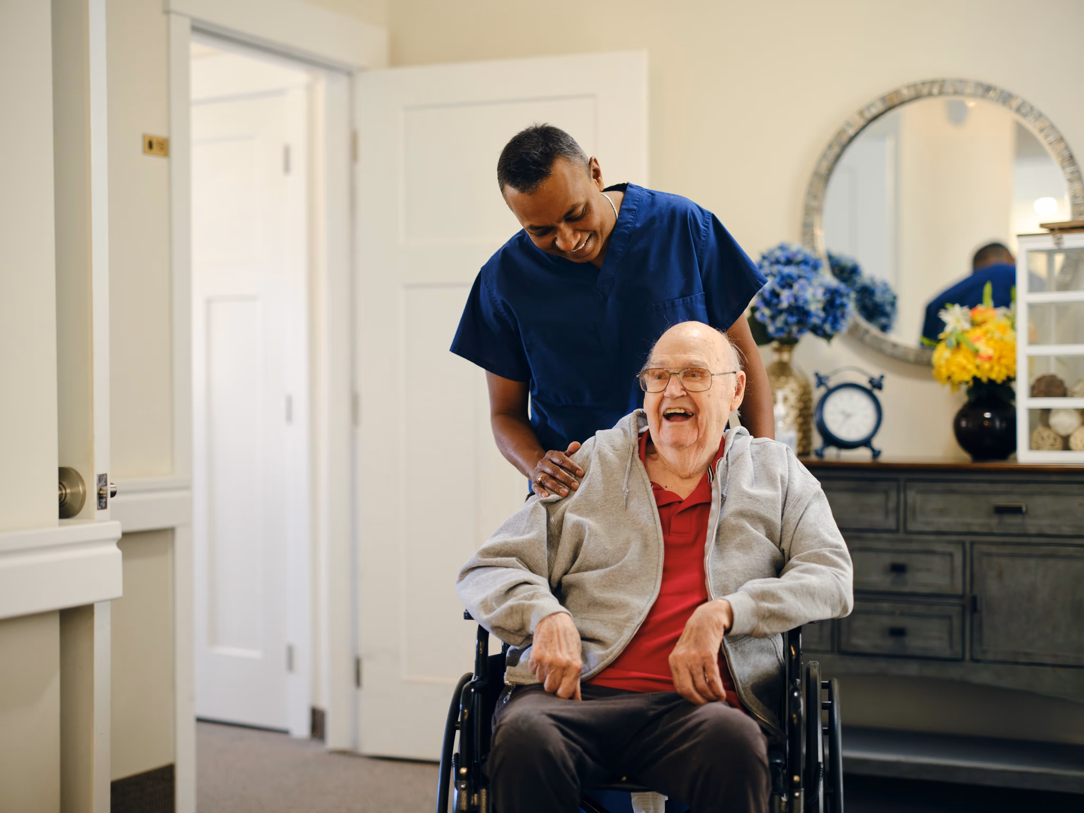 Smiling nurse in blue scrubs standing behind a happy elderly man in a wheelchair in a cozy room with flowers and a clock.