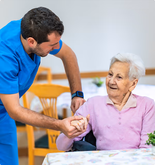 Healthcare worker in blue scrubs holding hands and smiling at an elderly woman seated at a table.