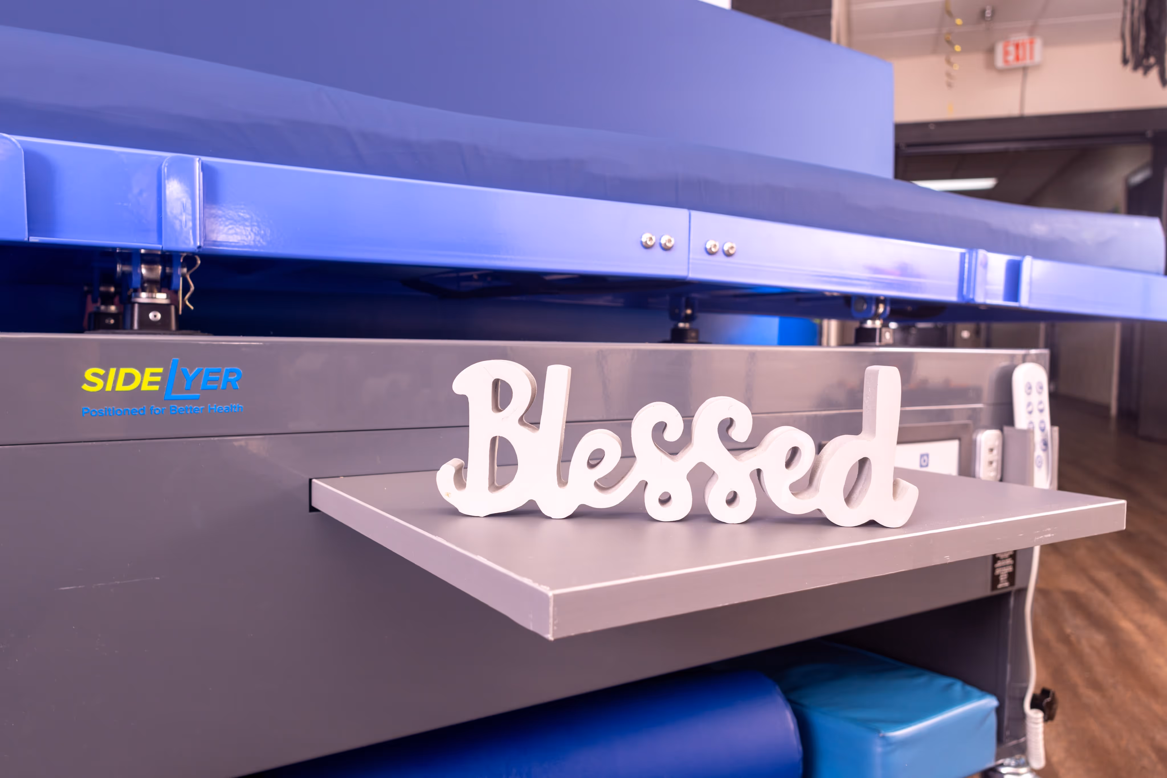 White decorative wooden word 'Blessed' displayed on a gray shelf attached to a blue and gray medical or therapy bed.