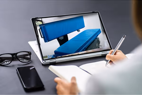 Person writing in a notebook in front of a tablet showing an image of blue medical beds, with glasses and a smartphone on the table.