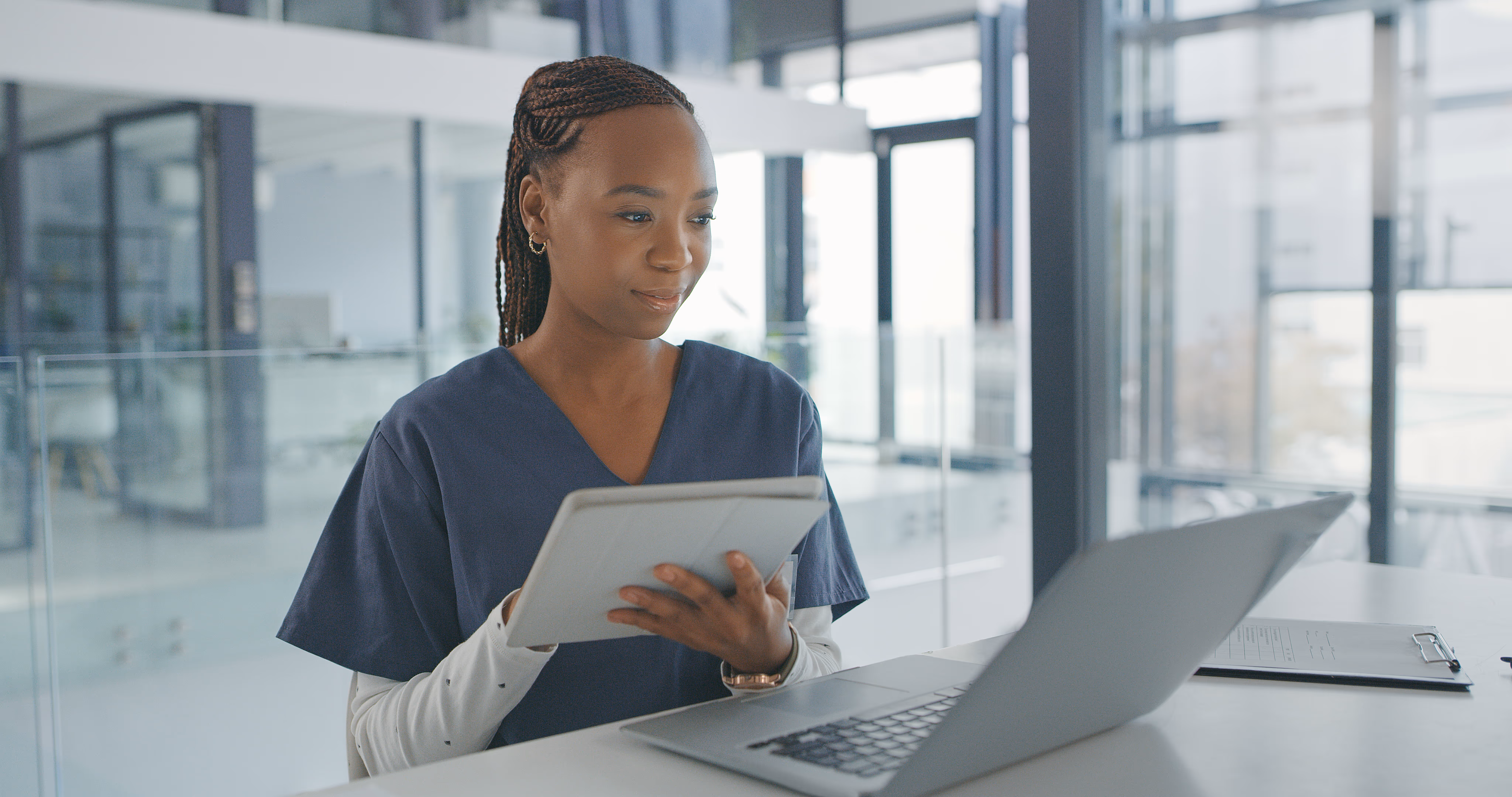 Female healthcare professional in scrubs holding a tablet and working on a laptop in a modern office.