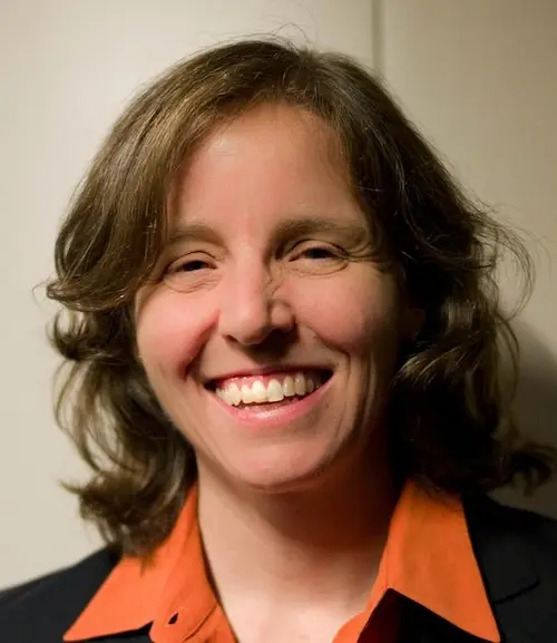 Smiling woman with shoulder-length brown hair wearing an orange collared shirt and dark jacket.