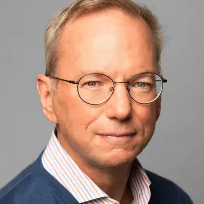 Portrait of an older man with short light hair wearing round glasses, a blue sweater, and a striped shirt against a gray background.