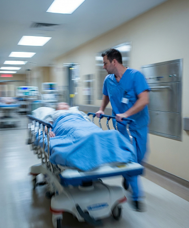 Healthcare worker in blue scrubs pushing a patient on a stretcher through a hospital hallway.