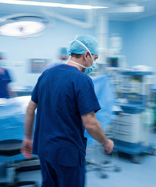 Surgeon in blue scrubs and surgical cap walking quickly through an operating room.