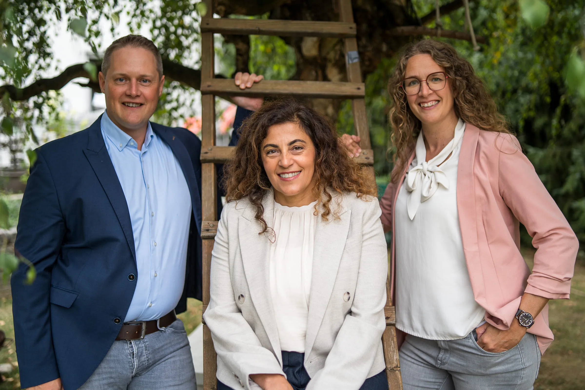 Drei lächelnde Personen vor einer Holzleiter, umgeben von Bäumen im Freien.