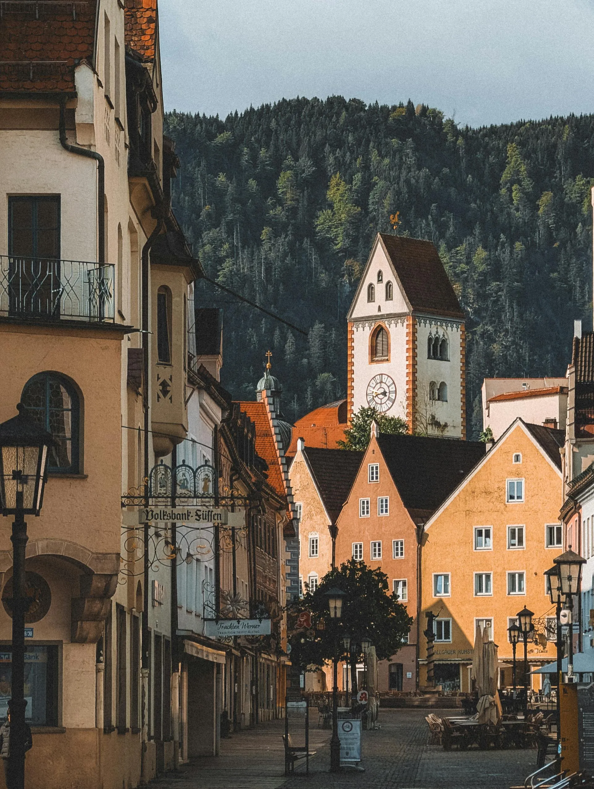 Historic street with yellow buildings, clock tower, outdoor café tables, and forested hill backdrop.