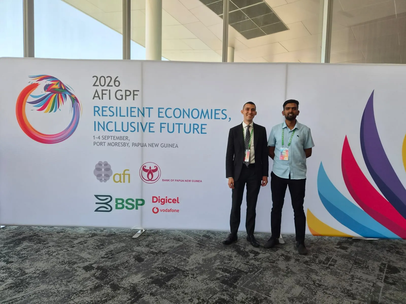 two smiling men standing against the poster of the event