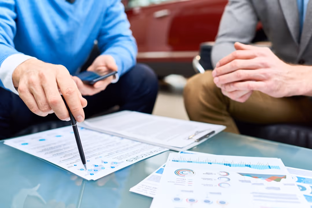 Two business professionals reviewing documents and data charts at a glass table, one pointing with a pen and holding a smartphone.